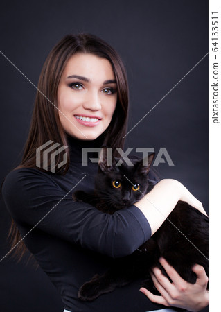 young brunette woman in black clothes on a black background with a black cat 64133511