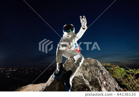 Astronaut sitting on rocky mountain and showing rock symbol. 64136110