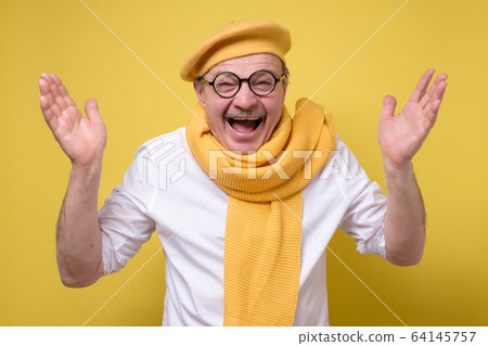 Surprised senior man in yellow scarf and beret holding hands up and smiling 64145757