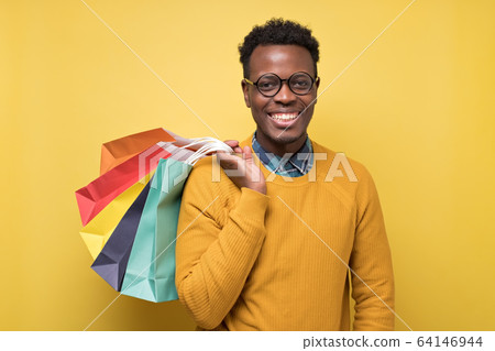 Young african american man in yellow sweater and glasses holding shopping bag Young african american man in yellow sweater and glasses holding shopping bag 64146944