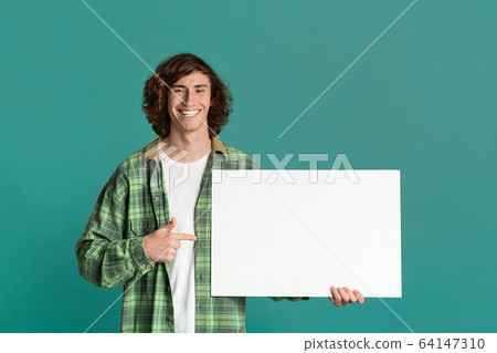 Joyful millennial guy pointing at blank poster on color background, space for your design 64147310