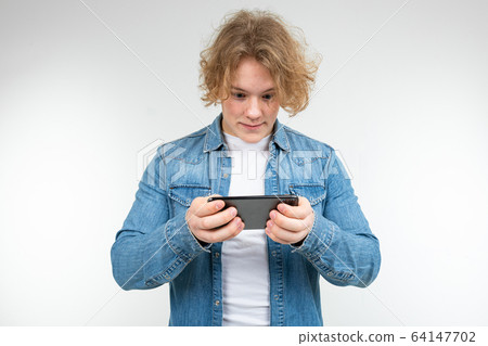 male gamer with blond hair in a denim jacket carefully plays on a smartphone on a white background 64147702