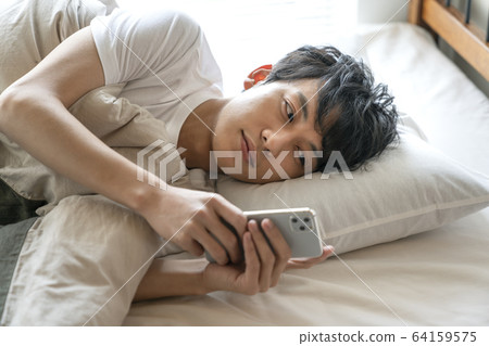 Young man looking at smartphone while sleeping in bed 64159575