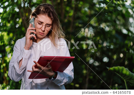 Young female agricultural engineer makes a call in greenhouse. Young female agricultural engineer makes a call in greenhouse. 64165078