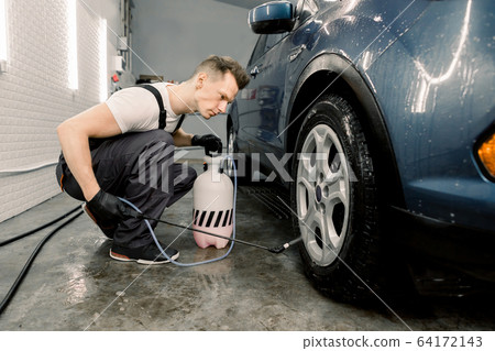 Shot of young Caucasian man in working clothes, car wash employee, washing the car dirty rim under high pressure water at professional auto washing service. Washing dirty car wheels with high pressure 64172143