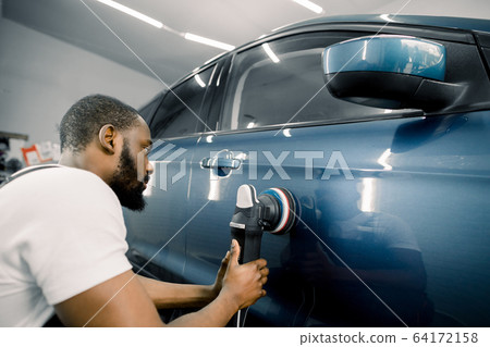 Young African man working at car detailing service, polishing blue car with polisher to eliminate contaminants from the surface of the car 64172158