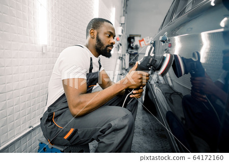 Auto detailing service, polishing of the car. Side view of young African American man worker n t-shirt and overalls, polishing blue car door with orbital polisher 64172160