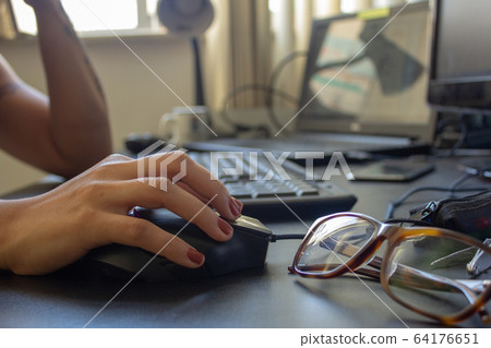 Closeup on the hand of a woman using her computer and glasses, with a keyboard, laptop and monitor on the background Closeup on the hand of a woman using her computer and glasses, with a keyboard, laptop and monitor on the background 64176651