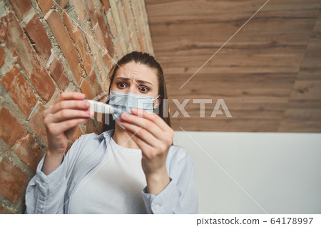 Ill female in a mask standing at the wall 64178997