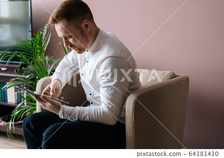 Young man sitting in soft armchair and using tablet computer at home. Young man sitting in soft armchair and using tablet computer at home. 64181410