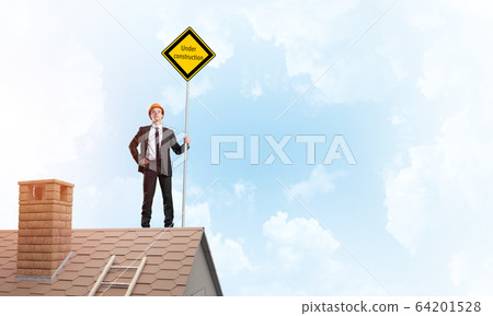 Young businessman on house brick roof holding yellow signboard. Mixed media Young businessman on house brick roof holding yellow signboard. Mixed media 64201528