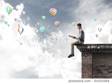 Man on roof edge reading book and aerostats flying in sky Man on roof edge reading book and aerostats flying in sky 64202068