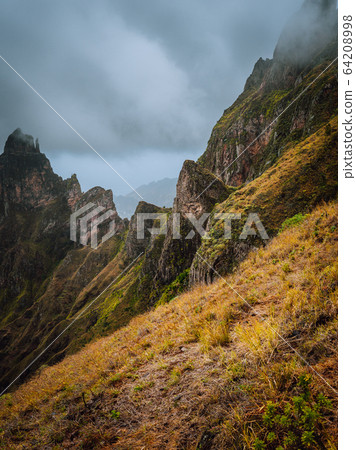 Moody view of Xo-Xo Valley, Santo Antao Island, Cape Verde Cabo Verde. Rugged mountain peak overgrown with verdant grass and encase by the fog. Some plants growing in foreground Moody view of Xo-Xo Valley, Santo Antao Island, Cape Verde Cabo Verde. Rugged mountain peak overgrown with verdant grass and encase by the fog. Some plants growing in foreground 64208998