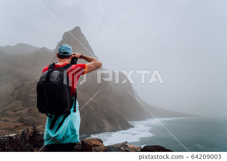 Male tourist with backpack on the scenic coastal road. The route leads along huge volcanic rock cliffs above roaring ocean and joins the towns of Cruzinha and Ponta do Sol. Santo Antao. Cape Verde Male tourist with backpack on the scenic coastal road. The route leads along huge volcanic rock cliffs above roaring ocean and joins the towns of Cruzinha and Ponta do Sol. Santo Antao. Cape Verde 64209003
