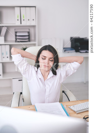 Young serene female office manager relaxing in armchair by desk Young serene female office manager relaxing in armchair by desk 64217590