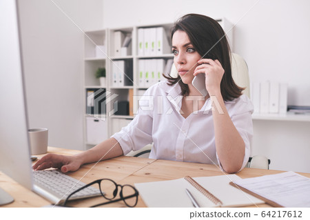Serious young secretary or office manager sitting by desk in front of computer 64217612