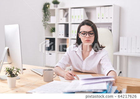Young serious businesswoman looking at financial paper while making notes 64217626