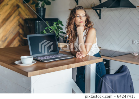 Pensive woman in the kitchen at the table with a laptop Pensive woman in the kitchen at the table with a laptop 64247413