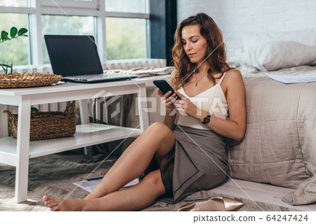 Woman in the bedroom is sitting on the floor and looking at the smartphone screen 64247424