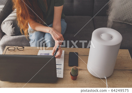 Woman freelancer uses a household humidifier in the workplace at home office with a laptop and documents. 64249575