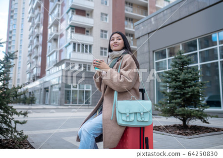 Dark-haired young woman in a beige coat having coffee Dark-haired young woman in a beige coat having coffee 64250830