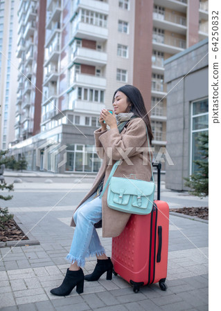Dark-haired young woman in a beige coat sitting on her suitcase 64250832