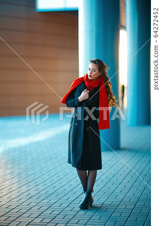 Young beautiful girl with perfect make-up, red lips, wearing a dark coat, red shirt, posing near shopping center. She just got out of high-end clothing store Young beautiful girl with perfect make-up, red lips, wearing a dark coat, red shirt, posing near shopping center. She just got out of high-end clothing store 64272452