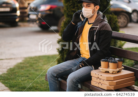 Courier in protective mask and medical gloves delivers pizza and coffe. Delivery boy sit on bench call to customer. Delivery service under quarantine. Coronavirus covid-19 theme Courier in protective mask and medical gloves delivers pizza and coffe. Delivery boy sit on bench call to customer. Delivery service under quarantine. Coronavirus covid-19 theme 64274539