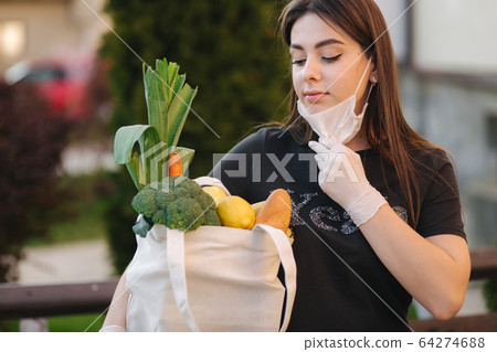 Young woman in mask and gloves hold bag with fresh food, vegetables fruits and bread. Female after grocery. Protect herself. Coronavirus theme 64274688