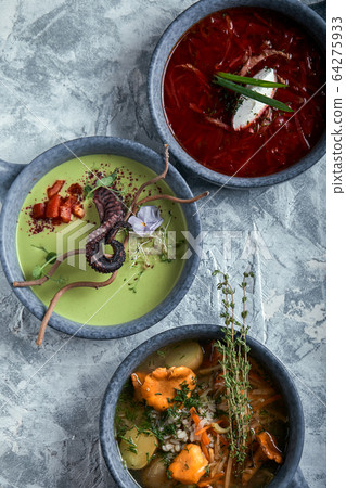 Gray plates with different soups on a gray background. Plate with asparagus soup with octopus, a plate of traditional borscht with sour cream, a plate with mushroom soup and standing on multi-colored 64275933