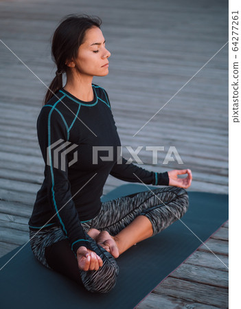 Concentrated girl in black practicing in yoga meditation on wooden seafront. Woman in lotus pose Concentrated girl in black practicing in yoga meditation on wooden seafront. Woman in lotus pose 64277261