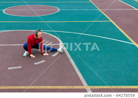 A young man in headphones stretches muscles before jogging on the sports ground early in the morning. A young man in headphones stretches muscles before jogging on the sports ground early in the morning. 64278559