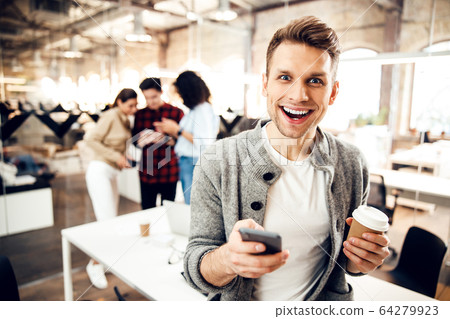 Cheerful young man holding smartphone and cup of coffee 64279923