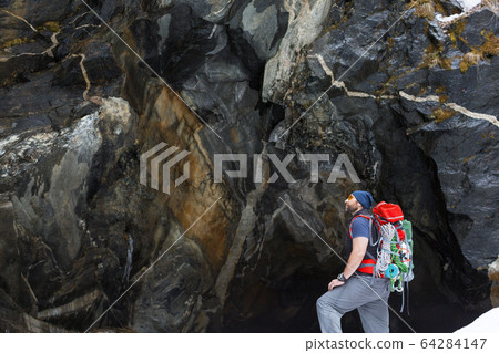 A man tourist with a backpack stands near a sheer stone rock. 64284147