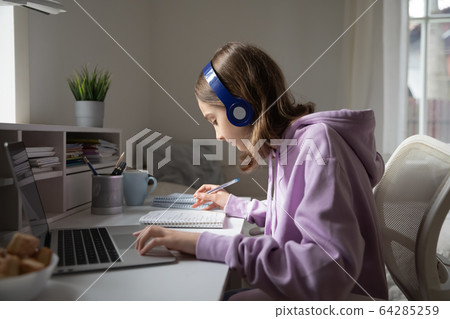 Teen girl wearing headphones studying online from home making notes Teen girl wearing headphones studying online from home making notes 64285259
