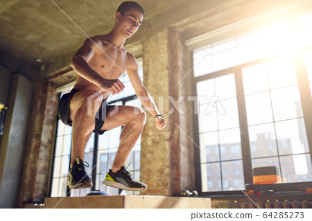Side view of jumping athlete man on wooden box in gym against background of large window 64285273