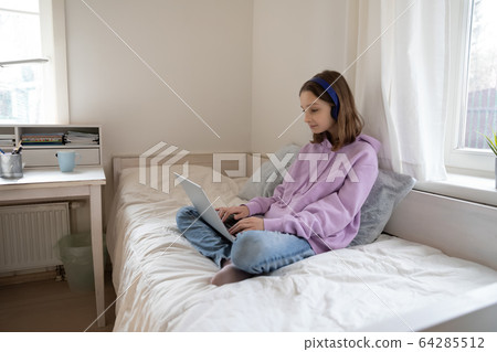 Teenage girl sitting on bed wearing headphones using laptop 64285512