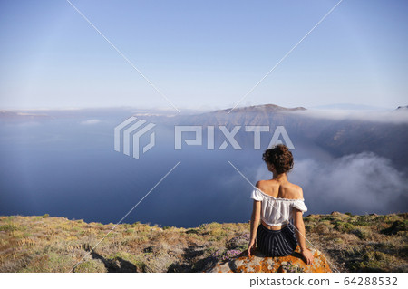 A young woman sits on the edge of a mountain and enjoys the view A young woman sits on the edge of a mountain and enjoys the view 64288532