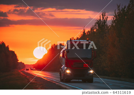 Red Truck Or Tractor Unit, Prime Mover, Traction Unit In Motion On Road, Freeway. Asphalt Motorway Highway Against Background Of Big Sunset Sun. Business Transportation And Trucking Industry. 64311619