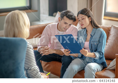 Dark-haired tall man sitting next to his girlfriend reading something 64314148