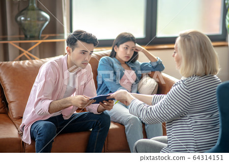 Dark-haired tall man sitting next to his girlfriend and passing documents to a blonde woman Dark-haired tall man sitting next to his girlfriend and passing documents to a blonde woman 64314151