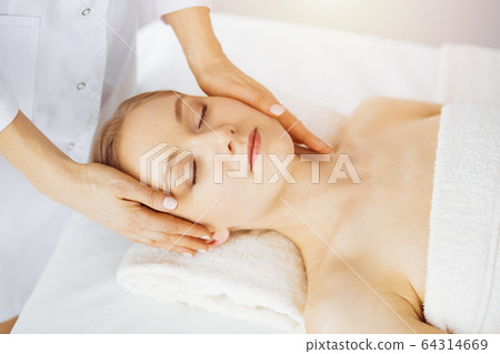 Beautiful caucasian woman enjoying facial massage with closed eyes in sunny spa salon. Relaxing treatment in medicine and Beauty concept 64314669