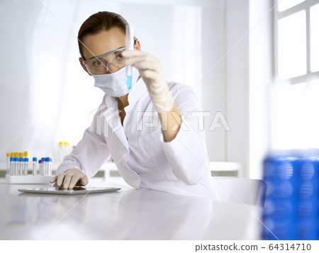 Female laboratory assistant in mask analyzing test tube with blue liquid. Medicine, health care and virus pandemic protection 64314710