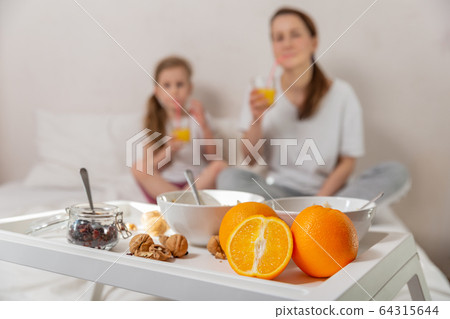 Happy mom and daughter have healthy breakfast on bed in a light bedroom on a sunny morning. A table Happy mom and daughter have healthy breakfast on bed in a light bedroom on a sunny morning. A table 64315644