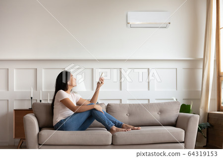 Vietnamese woman switching on air conditioner while resting seated indoors Vietnamese woman switching on air conditioner while resting seated indoors 64319153
