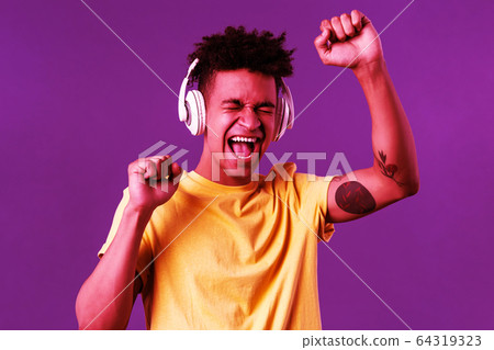 Close-up portrait of cheerful, happy young dancing guy lift hand up singing along, close eyes and smiling upbeat as listening awesome song in headphones, enjoying music, purple background 64319323