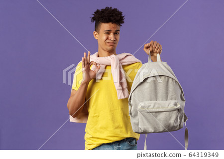 Good but not the best. Portrait of disappointed, unsatisfied young teenage student, guy grimacing without enthusiasm show okay, received new ugly backpack, standing purple background 64319349