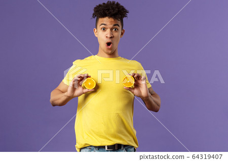 Holidays, vitamins and vacation concept. Portrait of funny and cute young 25s man fool around, showing breast with pieces of oranges over chest, look ashamed or shocked, purple background Holidays, vitamins and vacation concept. Portrait of funny and cute young 25s man fool around, showing breast with pieces of oranges over chest, look ashamed or shocked, purple background 64319407