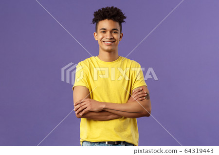 Close-up portrait of confident, smart and professional young male student with dreads, yellow t-shirt, cross arms over chest and smiling pleased, knows what he doing, purple background 64319443