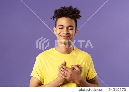 Always in my heart. Touched, carefree handsome young man with dreads, yellow t-shirt, close eyes and smiling, hold hand on chest as recall memory of his first love, feel heartwarming emotion 64319570